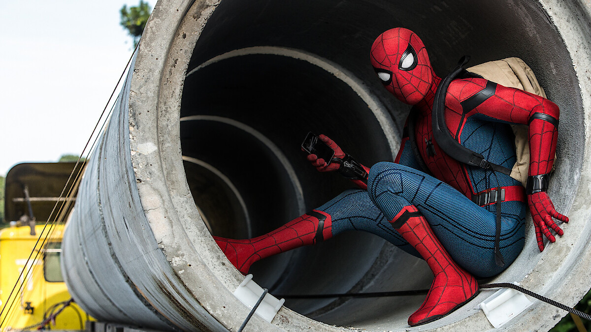A person in a Spider-Man costume crouches inside a large concrete pipe outdoors, holding a device and wearing a backpack, with construction vehicles and trees visible in the background.
