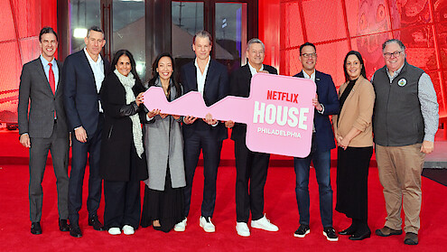 A group of people stand smiling on a red carpet in front of a building, holding a large pink key with "Netflix House Philadelphia" written on it, celebrating an event in a festive, welcoming atmosphere.