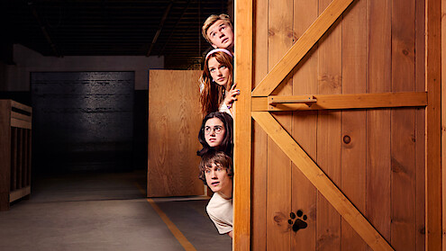 Four young people peeking around a wooden door with a paw print, in a dimly lit warehouse or storage environment, looking curious or cautious.