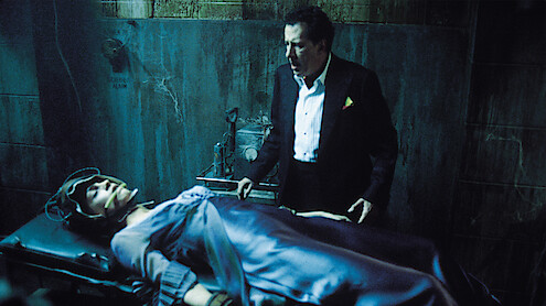 A man in a suit stands in a dimly lit, rundown room next to a restrained person lying on a table, creating a tense and dramatic atmosphere suggestive of a suspense or horror setting.