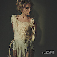 Woman with wavy blonde hair in an artistic, fringed cream top stands against a dark, plain background, looking to the side; moody editorial photo with dramatic lighting. Tudum Magazine logo in the corner.