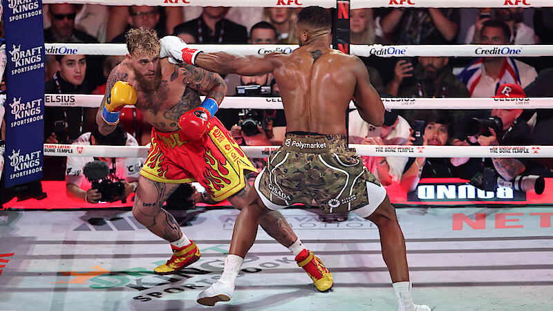 Jake Paul and Anthony Joshua fight in a brightly lit boxing ring, surrounded by ropes and an audience, with camera crews and sponsors visible in the background.