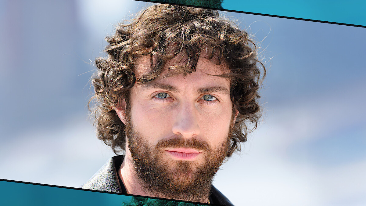 A man with curly brown hair and a beard is outdoors, wearing a black jacket, with a soft blurred background of blue sky and possibly distant mountains.