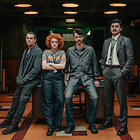 Four people in casual and business attire stand in a dimly lit office with wood paneling, retro décor, tiled floor, and an EXIT sign above a doorway, conveying a detective or investigative setting.