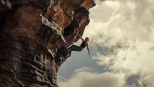 Person rock climbing on a steep, overhanging cliff with dramatic rock formations, reaching outward against a cloudy sky, outdoors in a rugged natural environment.