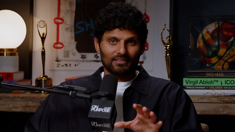 Man with curly hair speaks into a microphone labeled "On Purpose" in a cozy studio with books, awards, a lamp, and wall art in the background, suggesting a podcast or interview setting.