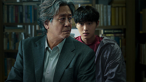 Two men, one older with gray hair in a suit and one younger with dark hair in casual clothes, are in a library or study filled with books, both appearing serious and focused.