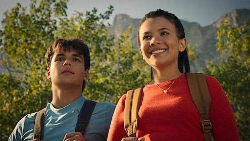 Two young people with backpacks, standing outdoors with trees and mountains in the background, enjoying a sunny day.