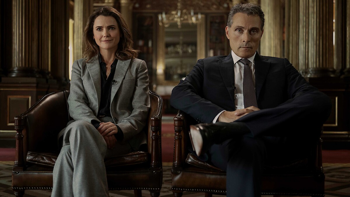 Two people in business attire sitting in leather chairs inside an opulent, historic room with gold columns and elegant decor, facing forward. The setting suggests a formal or professional environment.