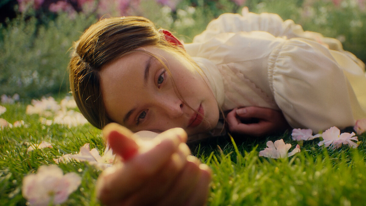 A young woman in a light blouse lies on green grass surrounded by pink flowers, looking directly at the camera in a sunlit garden or field.