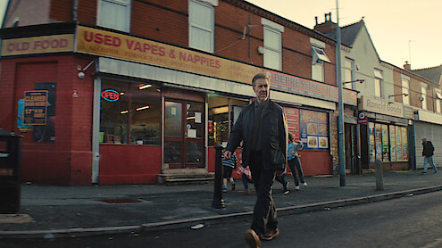 A man walks along a city street in front of a row of shops including a store selling used vapes; other people and children are seen on the sidewalk in a typical urban neighborhood setting, at dusk.