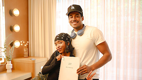 Two people smiling in a warmly lit room with vanity lights, one holding a script and making a peace sign, the other with headphones around his neck and wearing a baseball cap. Cozy indoor setting with a plant on a table.
