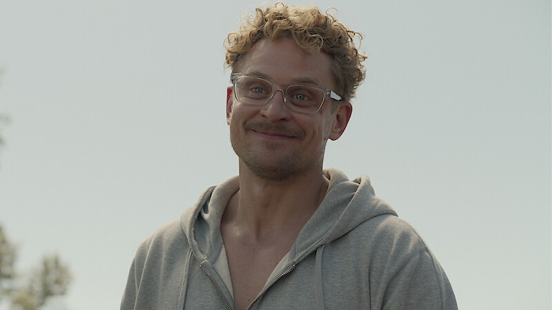 A smiling man with curly hair and glasses wearing a gray hoodie stands outdoors under a clear sky, with some trees visible in the background.