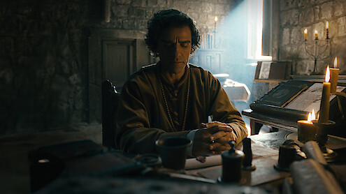 A man sits at a medieval wooden desk in a dimly lit stone room, surrounded by candles, books, and writing tools, with sunlight streaming through a small window, creating a contemplative atmosphere.
