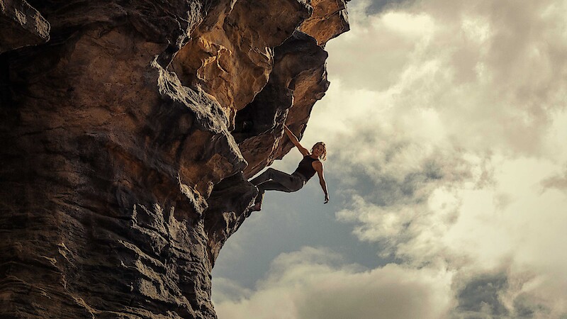 A rock climber scaling a steep, overhanging cliff outdoors under a cloudy sky, demonstrating strength and balance in a dramatic natural setting.