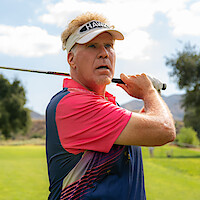 Man in a pink and blue polo shirt and visor holding a golf club in a follow-through pose on a sunny golf course with green grass, trees, and hills in background.