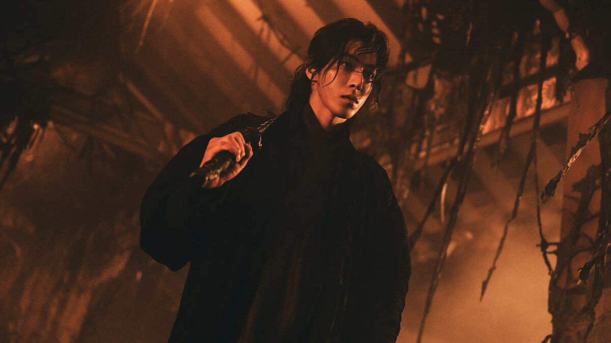 Person in dark clothing holding a weapon, standing in a dimly lit, eerie environment with hanging vines and dramatic orange lighting, evoking a tense, mysterious atmosphere.