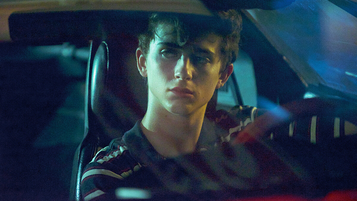 Young man sitting in the driver's seat of a car at night, looking out the windshield with a serious expression, illuminated by dim, moody blue and yellow lighting inside the vehicle.
