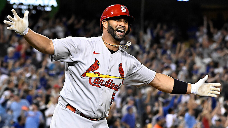 Albert Pujols in a St. Louis Cardinals uniform celebrating on field with arms outstretched, smiling, with cheering crowd in stadium background during night game.
