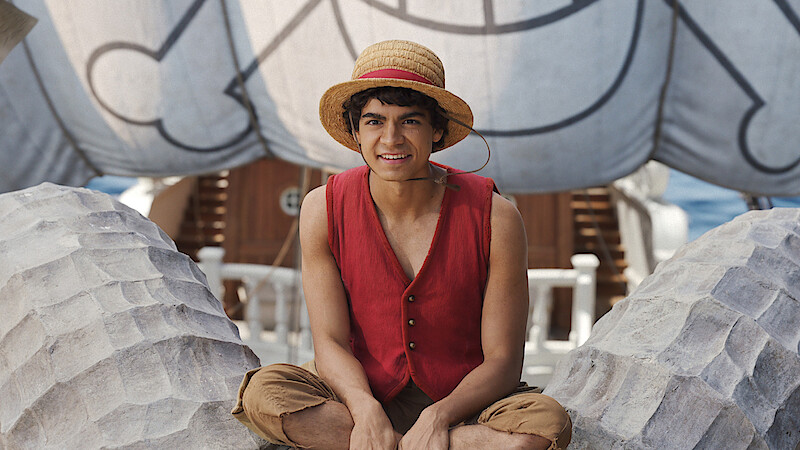 Luffy, a young man in a straw hat and red vest sits cross-legged on a ship's deck, with large decorative sails and nautical details in the background, suggesting a lively maritime adventure setting.