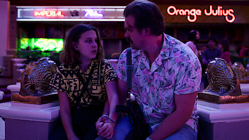 A man and a teenage girl sit closely on a mall bench, surrounded by neon lights and storefronts.