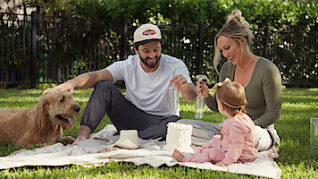 Baker Mayfield with his family and dog enjoying a picnic on a blanket in a sunny backyard, playing with objects and smiling, surrounded by greenery and a fence in the background.