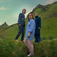 Three people stand on grassy hills under a cloudy sky, dressed in modern, colorful clothes, with a mountain peak in the background, creating a contrast between natural landscape and urban fashion.