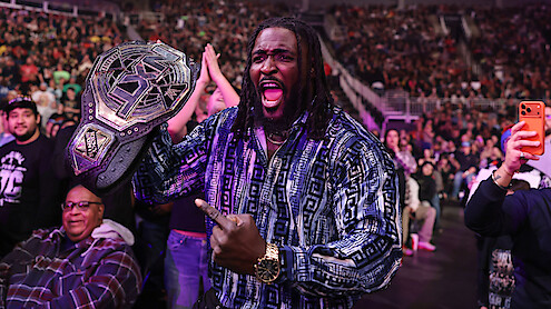Oba Femi holding a wrestling championship belt and celebrating in a crowded arena, surrounded by fans taking photos and cheering during a live event.
