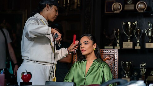 A woman in a green blouse sits in a carved chair while a stylist adjusts her hair. They are in a room with a display of trophies and awards on shelves in the background.