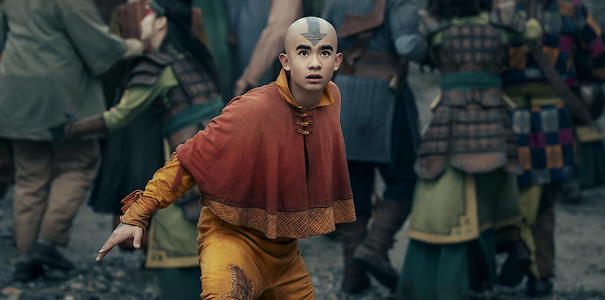 A young person in orange and yellow monk-style clothing with a blue arrow tattoo on their head stands alert in a crowded outdoor setting with people in armor, suggesting a tense or urgent situation.
