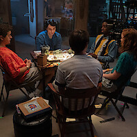 Five teenagers sitting around a table playing a board game in a dimly lit basement, surrounded by various objects and filming equipment, creating a cozy and nostalgic atmosphere.