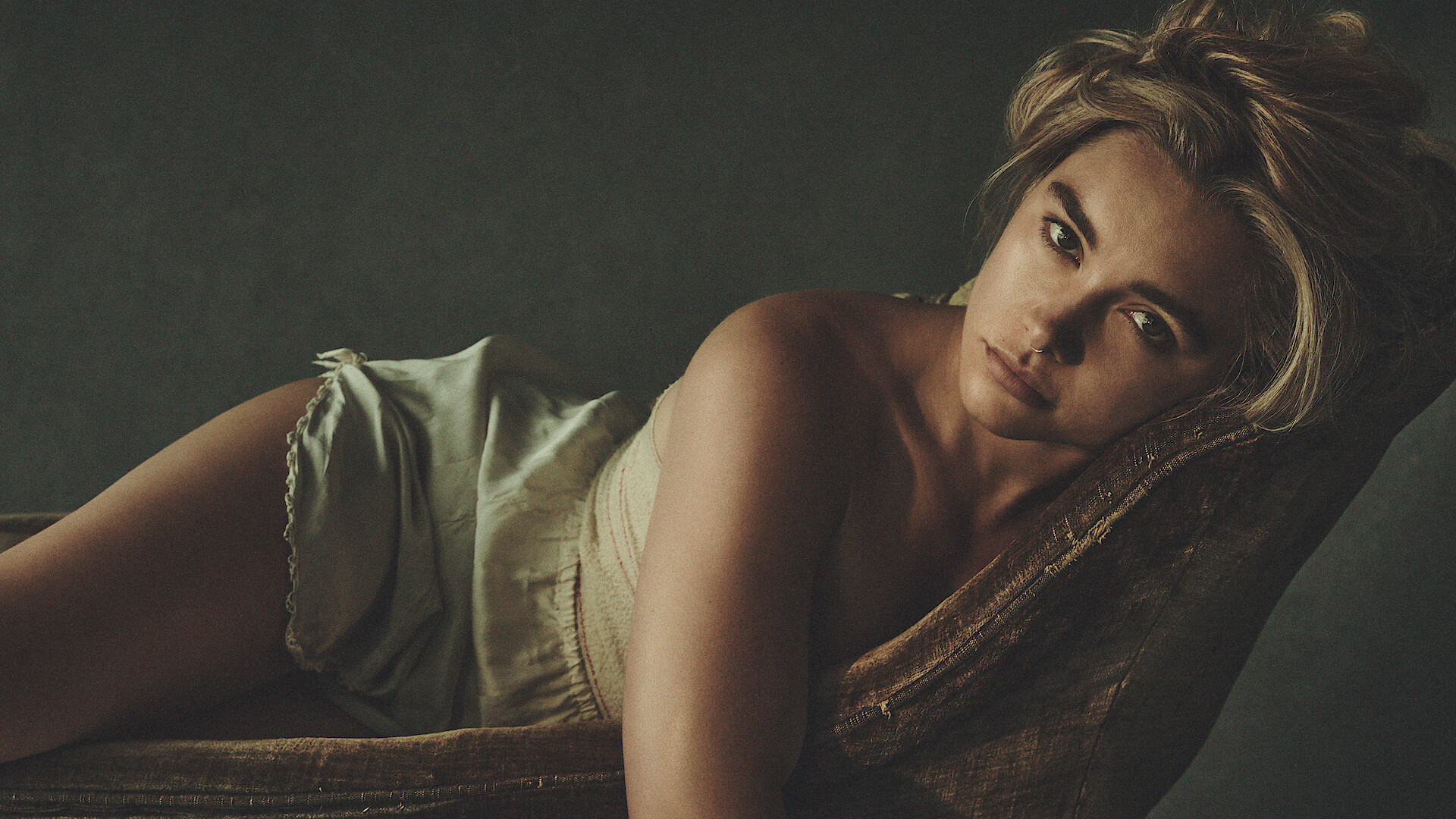 Woman lying sideways on a vintage chair, wearing light sleepwear, looking directly at the camera in a softly lit, moody indoor setting with a dark background.