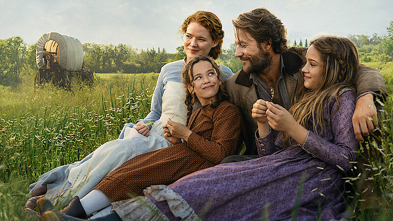 A family of four in historical clothing sits in a grassy field, smiling and enjoying time together, with a covered wagon and trees in the background on a sunny day.