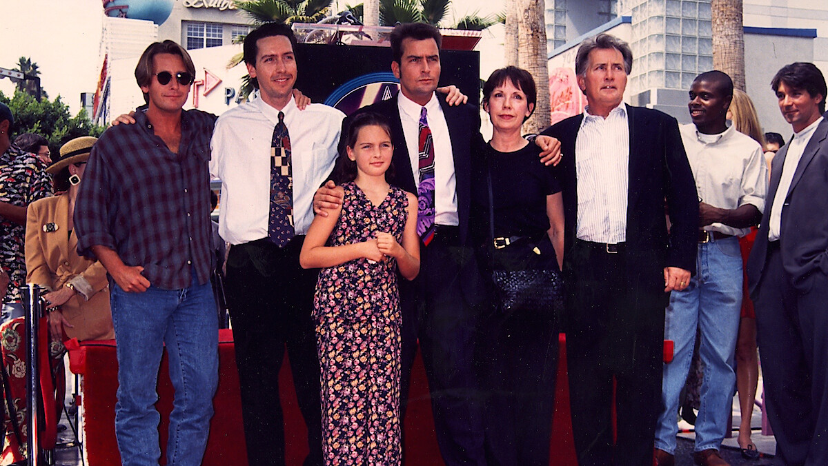 A group of people posing together outdoors on a red carpet, dressed in formal and casual attire, with palm trees and buildings in the background during a sunny day event.