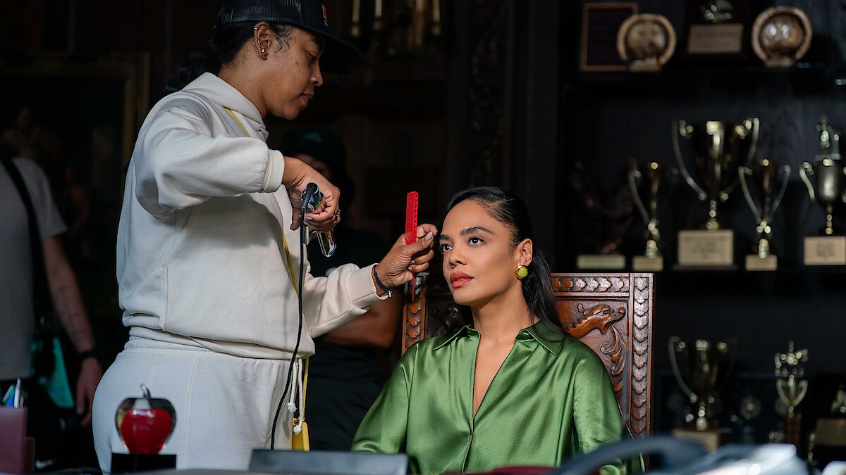 A woman in a green blouse sits in a carved chair while a stylist adjusts her hair. They are in a room with a display of trophies and awards on shelves in the background.