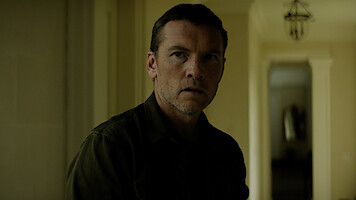 A serious man stands indoors in a dimly lit hallway with pale yellow walls and columns, looking slightly off-camera. The background features a corridor and a hanging light fixture.