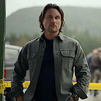 A man with medium-length hair and a serious expression stands outdoors near yellow caution tape, with a forested mountain and blurry people in the background, suggesting a rural or investigative setting.