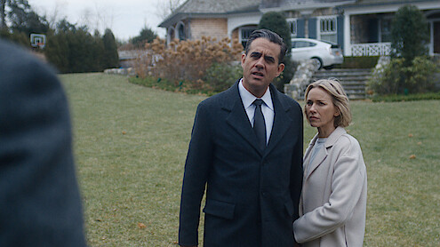 Man in dark coat and woman in light coat stand together on a lawn, looking concerned, in front of a large house with stone steps and autumn landscaping.