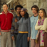 Five young adults standing close together outdoors by a stone wall, looking concerned or surprised. One person in the center holds a phone. They are dressed in casual, colorful clothing, suggesting a contemporary urban or historical setting.