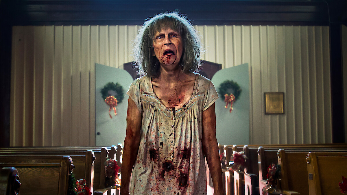 A disheveled, bloodied woman with gray hair stands in the aisle of a decorated church, with pews on either side and wreaths on the doors in the background, looking distressed or possibly zombified.