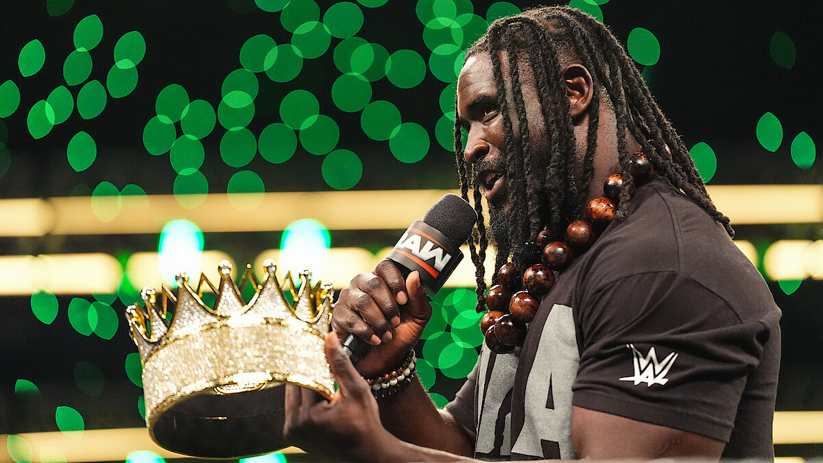 Wrestler with long braids and beaded necklace speaks into a microphone labeled "RAW" while holding a large golden crown, standing in a brightly lit arena with green lights in the background.