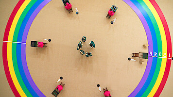 Overhead view of people in colorful uniforms inside a bright arena with rainbow circles, numbered blocks, and a game-like atmosphere, creating a tense yet playful mood.