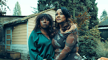 Two women stand close together outside beside a rustic shed, surrounded by greenery and trees. Both look thoughtful, dressed formally, with overcast natural lighting creating a serene, introspective mood.