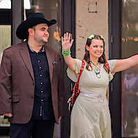 Man in a black cowboy hat and brown blazer stands next to a smiling woman in a light dress with necklaces and flower hair clips, waving, outside a building with glass doors.
