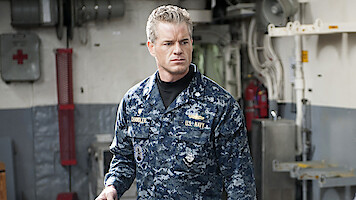 A man in a blue digital camouflage Navy uniform stands indoors on a ship, surrounded by industrial equipment, pipes, and emergency gear, looking serious and focused.