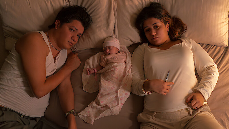A man, woman, and newborn baby lie in bed together, parents on either side of the baby who is swaddled and wearing a hat. The mood is subdued, with soft, warm lighting suggesting nighttime or early morning at home.