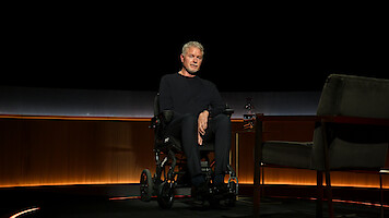 Eric Dane sits in a wheelchair on a dimly lit stage next to an empty chair and a small table with a glass carafe, in a modern studio interview setting with warm lighting and dark background.