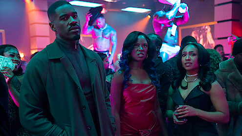 Three people in stylish outfits stand in a crowded nightclub with neon lighting; shirtless dancers perform on stage in the background, and audience members watch and hold money.
