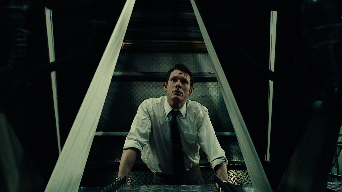 Man in white shirt and tie looks upward in a dimly lit industrial or mechanical setting, framed by metallic structures, suggesting a tense or suspenseful atmosphere.