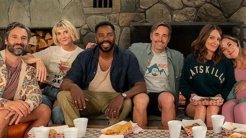 Six adults sitting close together on a couch in a cozy room with a stone fireplace, enjoying a casual meal with trays of food and drinks, smiling and relaxed in a friendly, informal setting.